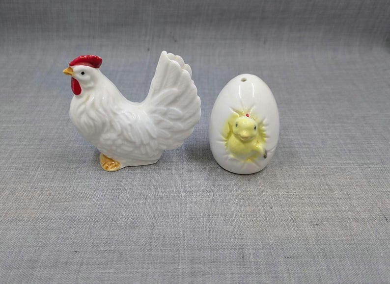 Puede incluir: Una figura de gallina de cer&aacute;mica blanca con cresta roja y pico amarillo se encuentra junto a una figura en forma de huevo blanco con un pollito amarillo que emerge. Son objetos decorativos.