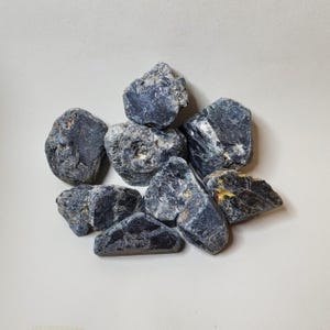 May include: A collection of rough, dark blue-grey gemstones with textured surfaces. The stones vary in size and shape, with some showing hints of lighter grey and brown. The arrangement is scattered against a plain, light background.