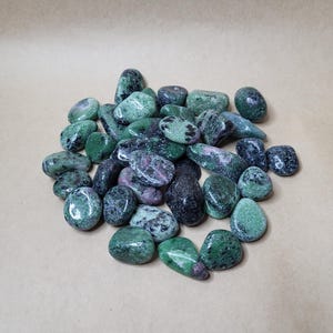 May include: A pile of polished ruby in zoisite stones. The stones are a mix of deep green and black, with pink and red inclusions. The gemstones are smooth and rounded, with a natural appearance. The stones vary in size and shape.