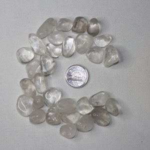 Peut inclure: Une collection de pierres de quartz clair roulées, disposées en demi-cercle autour d'une pièce de monnaie canadienne. Les pierres sont lisses et polies, avec une apparence translucide.