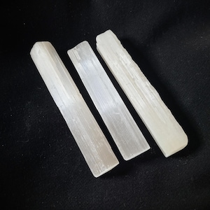 Peut inclure: Trois baguettes de cristal de sélénite blanches sont disposées en diagonale sur un fond noir. Les baguettes ont un aspect translucide et une forme rectangulaire lisse avec des bords naturels et irréguliers. Les cristaux sont de couleur blanc cassé.