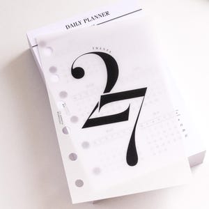 Può includere: Una pagina di ricarica per agenda giornaliera con il grande numero 27 stilizzato stampato in nero su un foglio di carta velina traslucida. La pagina presenta fori perforati per la rilegatura, il testo DAILY PLANNER e una matita dorata.