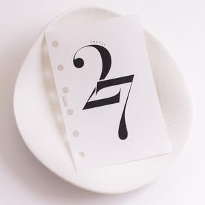 May include: A white planner insert page featuring a large, stylised black number 27 with the word TWENTY printed in small letters above it. The paper has a vertical row of six circular punch holes along the left edge and small text on the side.