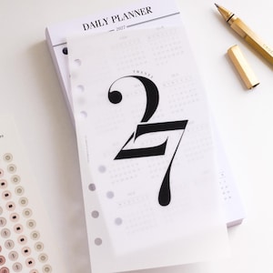 May include: A 2027 daily planner with a translucent vellum cover featuring the large black text 27. The planner has a white paper interior with printed text and a gold-coloured metal pen resting nearby, alongside a sheet of small circular stickers.