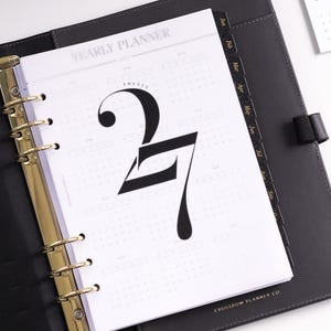 Peut inclure: Un classeur en cuir noir avec des anneaux dorés contenant un agenda annuel. La page affiche le texte YEARLY PLANNER et un grand 27 stylisé. Des onglets mensuels noirs sont visibles sur le côté, marqués des mois de Jan à Sep.