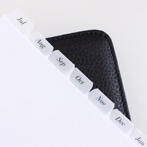 May include: A black leather planner featuring white translucent index tabs labeled with the months Jul, Aug, Sep, Oct, Nov, Dec, and Jan. The planner pages are white with black text, and the cover has a textured, pebbled surface finish.