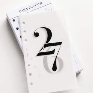 May include: A stack of white daily planner pages with a transparent overlay featuring the number 27 in a bold, elegant font. A gold pen and pen cap are placed on the white surface. The planner is labelled "DAILY PLANNER" and "2026-27".