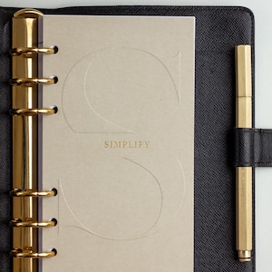 May include: A black leather binder with a gold six-ring mechanism and a matching gold pen. The beige insert features a large embossed letter S, the word SIMPLIFY in gold, and the text Less clutter, less stress, more focused impact.