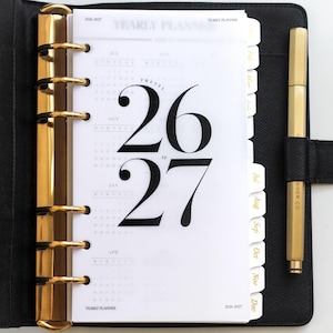 Peut inclure: Agenda en cuir noir avec des détails dorés, ouvert sur une page avec les chiffres "26 to 27" en gros caractères gras. Un stylo doré est posé à droite. L'agenda a des onglets mensuels.