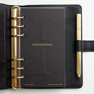 May include: A black leather planner with a gold six-ring binder mechanism and a matching gold pen. The black cover insert features a large embossed letter I, the word INTENTIONAL in gold, and the text Living on purpose and leaving nothing to default.