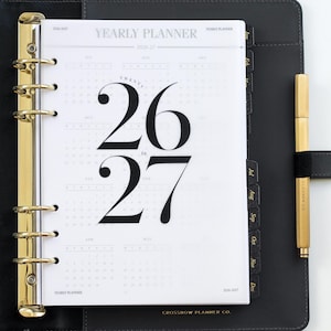 Peut inclure: Un agenda en cuir noir avec des détails dorés, ouvert sur une page avec le texte en gras "26 to 27". L'agenda comprend des onglets mensuels et un stylo doré. Le texte "YEARLY PLANNER" et "CROSSBOW PLANNER CO." sont visibles.