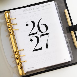 Peut inclure: Un agenda annuel avec un classeur doré et un stylo. L'agenda est ouvert sur une page avec les chiffres "26 27" en gros caractères gras. L'agenda comporte des onglets mensuels et le texte "YEARLY PLANNER" en haut.