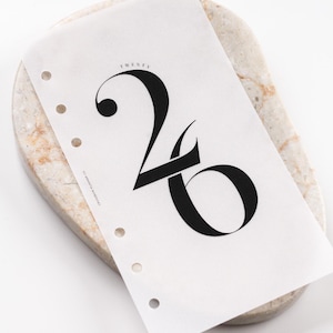 Peut inclure: Une page de planificateur blanche avec le chiffre "26" en gras noir. Le mot "TWENTY" est au-dessus du chiffre. La page est perforée pour la reliure et repose sur une pierre ovale de couleur claire.