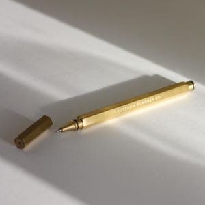 Könnte beinhalten: Ein goldfarbener Stift mit sechseckigem Schaft und passender Kappe, der auf einer weißen Oberfläche liegt. Der Stift ist mit "CROSSBOW PLANNER CO." beschriftet. Die Kappe ist abgenommen und gibt die Spitze des Stifts frei. Der Stift ist etwa 14 cm lang.