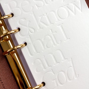 Könnte beinhalten: Eine weiße Planereinlage mit dem geprägten Text Be still, & know that I am God. Das Papier wird von einer goldfarbenen Sechs-Ring-Bindemechanik aus Metall gehalten, die an einem strukturierten braunen Einband am linken Rand befestigt ist.