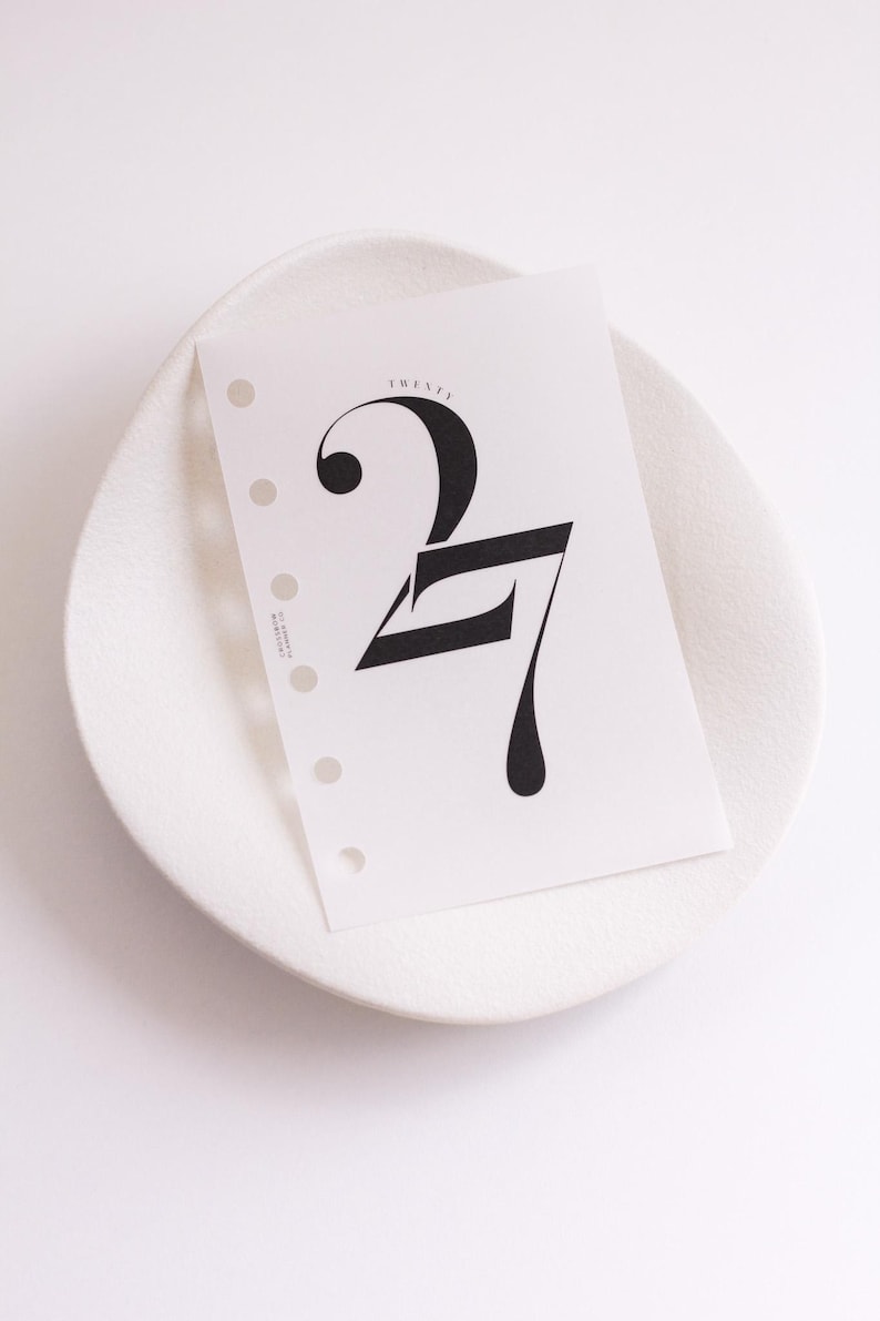 May include: A white planner insert page featuring a large, stylized black number 27 with the word TWENTY printed in small letters above it. The paper has a vertical row of six circular punch holes along the left edge and small text on the side.