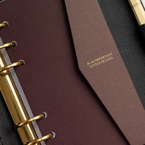 Könnte beinhalten: Ein dunkelbrauner Ringplaner mit goldfarbenen Metallringen und einer Klappe, die den goldgeprägten Text "for the IMPORTANT LITTLE DETAILS" trägt. Der Planer liegt auf einem schwarzen, strukturierten Hintergrund mit einem goldenen Stift.