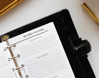 May include: A black leather-textured binder containing a monthly planner insert with white pages. The page features headers for Monthly Planner, Goals & Priorities, Must-Do Tasks, and Important Dates, with a gold ruler visible at the top.