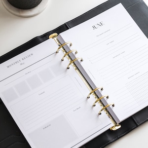 May include: A black leather six-ring binder planner open to pages labeled Monthly Review for May and June. The planner features gold-toned metal rings, white paper with black text, and sections for intentions, tasks, habits, and gratitude.