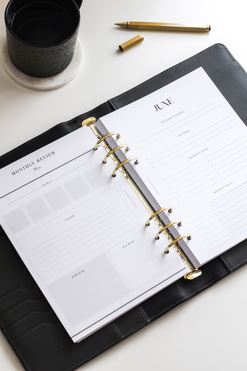 May include: A black leather six-ring binder planner open to pages labeled Monthly Review for May and June. The planner features gold-toned metal rings, white paper with black text, and sections for intentions, tasks, habits, and gratitude.