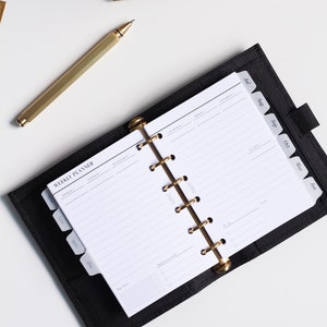 May include: A black leather weekly planner with a gold six-ring binder mechanism. The open planner displays white pages with printed text including "WEEKLY PLANNER" and days of the week. A gold pen lies diagonally next to the planner on a white surface.