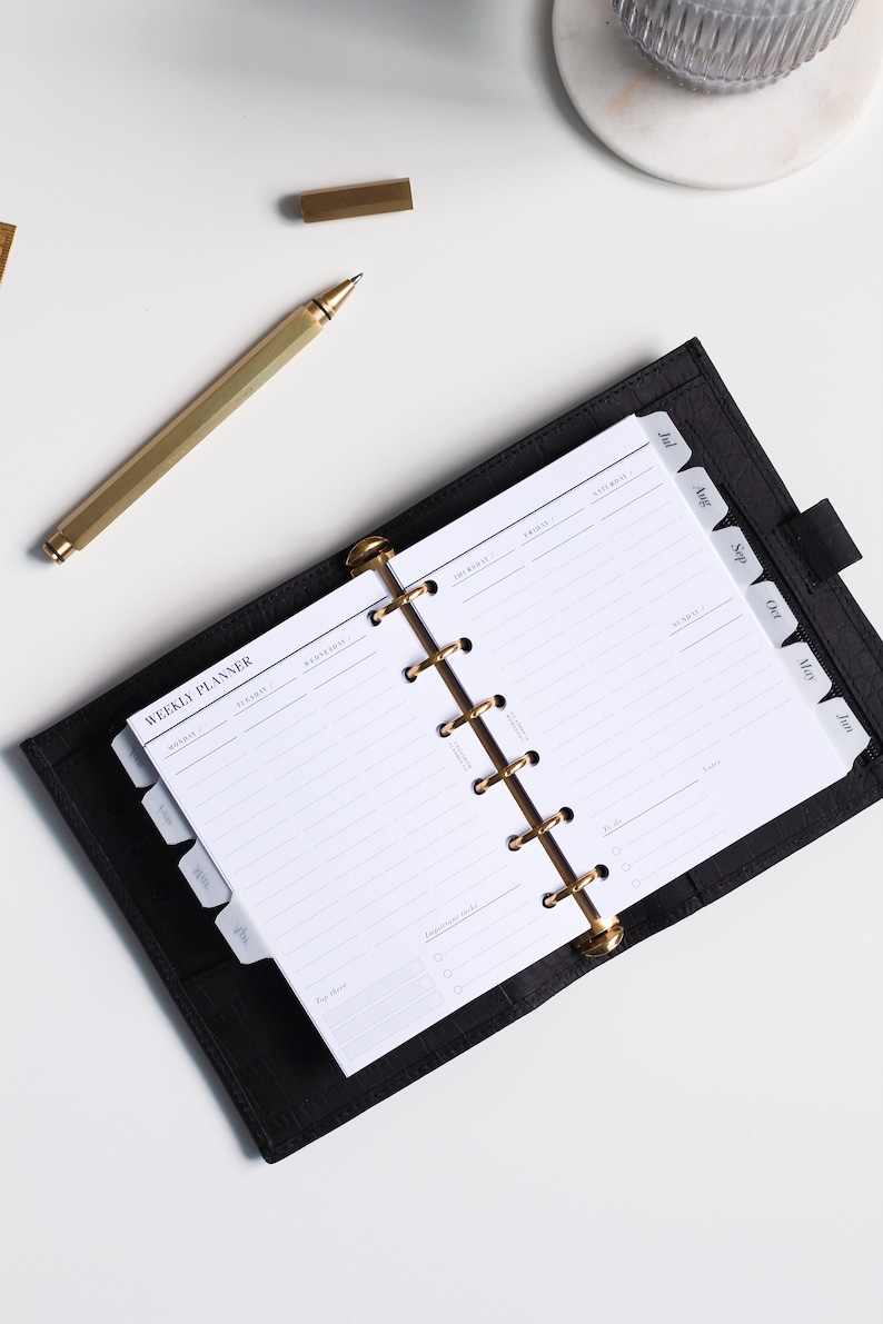 May include: A black leather weekly planner with a gold six-ring binder mechanism. The open planner displays white pages with printed text including "WEEKLY PLANNER" and days of the week. A gold pen lies diagonally next to the planner on a white surface.