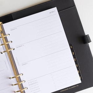 May include: A black leather-bound planner with a gold-toned six-ring binder mechanism. The white paper insert displays a daily schedule for Friday 30, Saturday 31, and Sunday 1, with the text June 1 and Crossbow Planner Co. printed on the page.