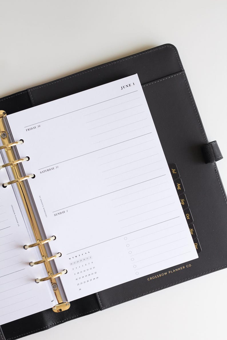 May include: A black leather-bound planner with a gold-toned six-ring binder mechanism. The white paper insert displays a daily schedule for Friday 30, Saturday 31, and Sunday 1, with the text June 1 and Crossbow Planner Co. printed on the page.