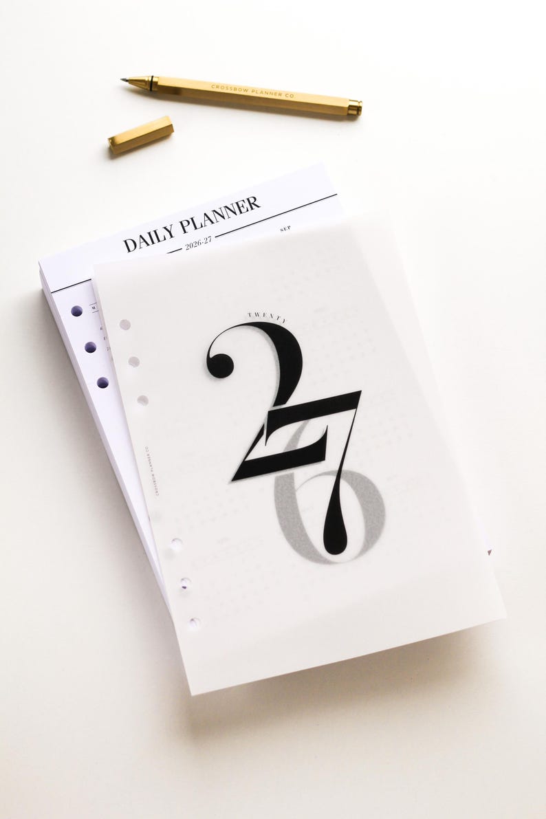 May include: A white daily planner with the text "DAILY PLANNER 2026-27" and a gold pen. The planner has a translucent overlay with the number "27" in a bold, stylized font. The pen is gold and has the text "CROSSTOWN PLANNER CO."