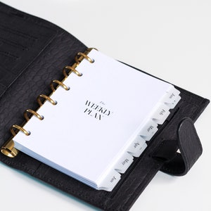 May include: A black leather-textured planner with a gold-toned ring binder mechanism. The white interior pages feature the text The WEEKLY PLAN and include monthly tab dividers labeled Jan, Feb, Mar, Apr, May, and Jun along the right edge.