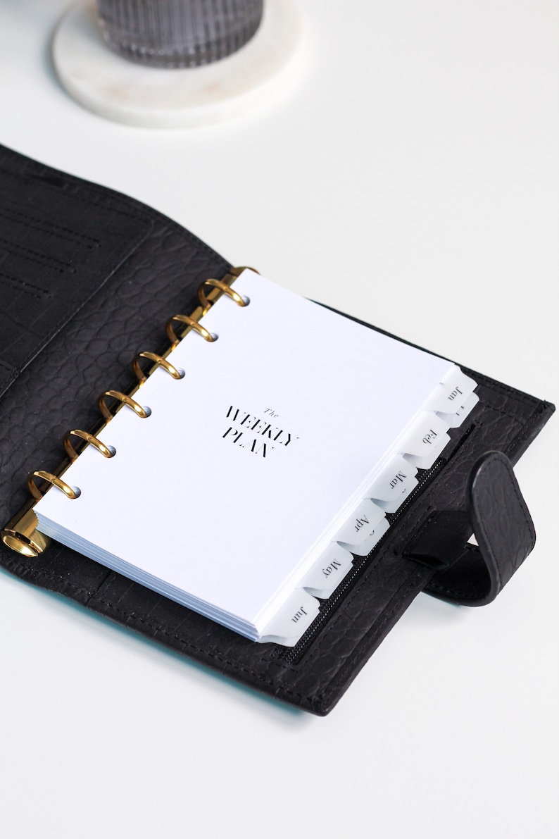 May include: A black leather-textured planner with a gold-toned ring binder mechanism. The white interior pages feature the text The WEEKLY PLAN and include monthly tab dividers labeled Jan, Feb, Mar, Apr, May, and Jun along the right edge.