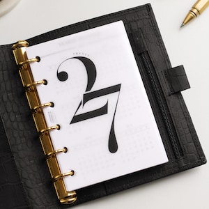 May include: A black leather planner with a crocodile texture, featuring gold-toned metal rings and a white insert page. The page displays the large black text '27' and the word 'TWENTY' above it, alongside a small calendar grid.