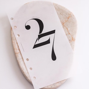 May include: A translucent planner insert page featuring the large black number 27 in a stylized serif font. The word TWENTY is printed in small letters above the number, and the text EPHEMERA PLANNER CO is visible on the left side near the punch holes.