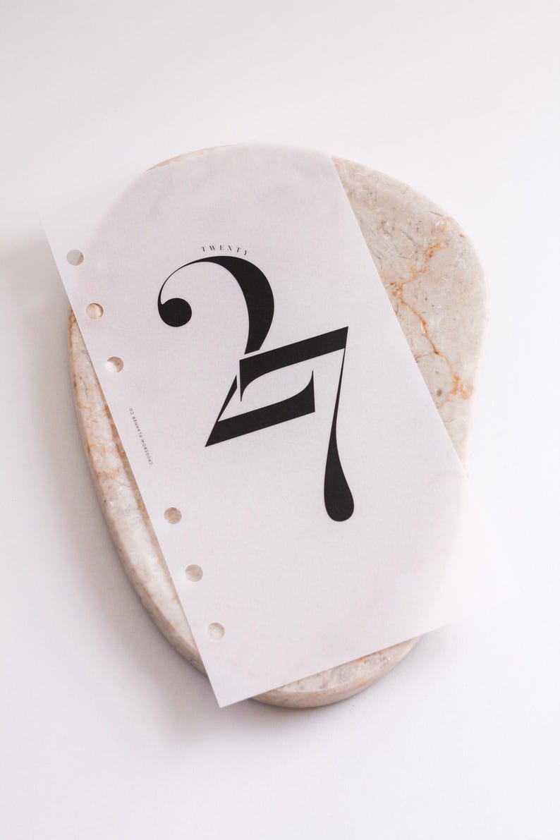 May include: A translucent planner insert page featuring the large black number 27 in a stylized serif font. The word TWENTY is printed in small letters above the number, and the text EPHEMERA PLANNER CO is visible on the left side near the punch holes.