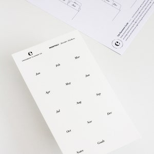 May include: A white sheet of monthly divider stickers featuring black text for each month from January to December, plus Notes and Goals. The sheet includes the Crossbow Planner Co. logo. A separate tab placement guide sheet is also visible.