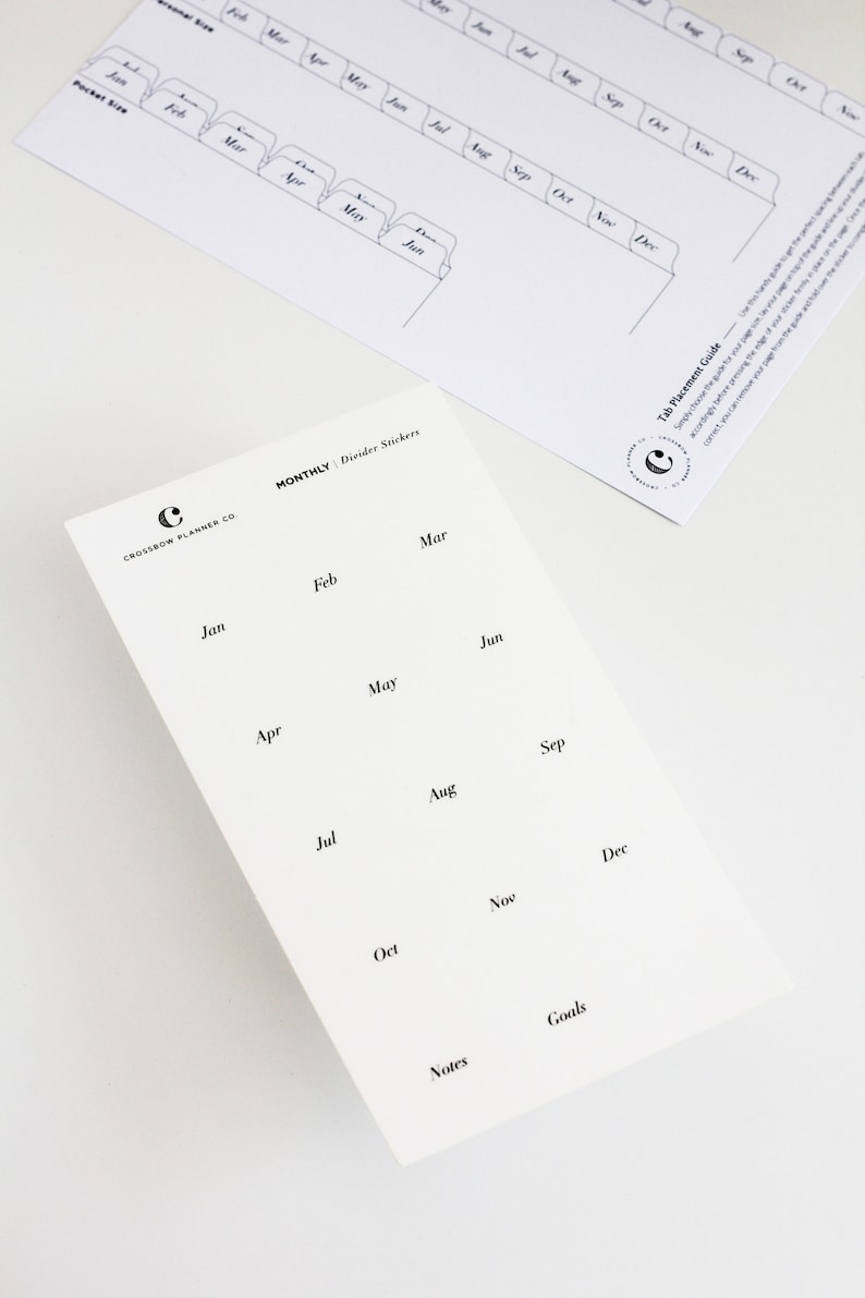 May include: A white sheet of monthly divider stickers featuring black text for each month from January to December, plus Notes and Goals. The sheet includes the Crossbow Planner Co. logo. A separate tab placement guide sheet is also visible.