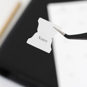 May include: A small white adhesive tab with a flared shape and the word Notes printed in a black serif font. The tab is held by a pair of black metal tweezers, showcasing its design for organization and labeling in planners or notebooks.