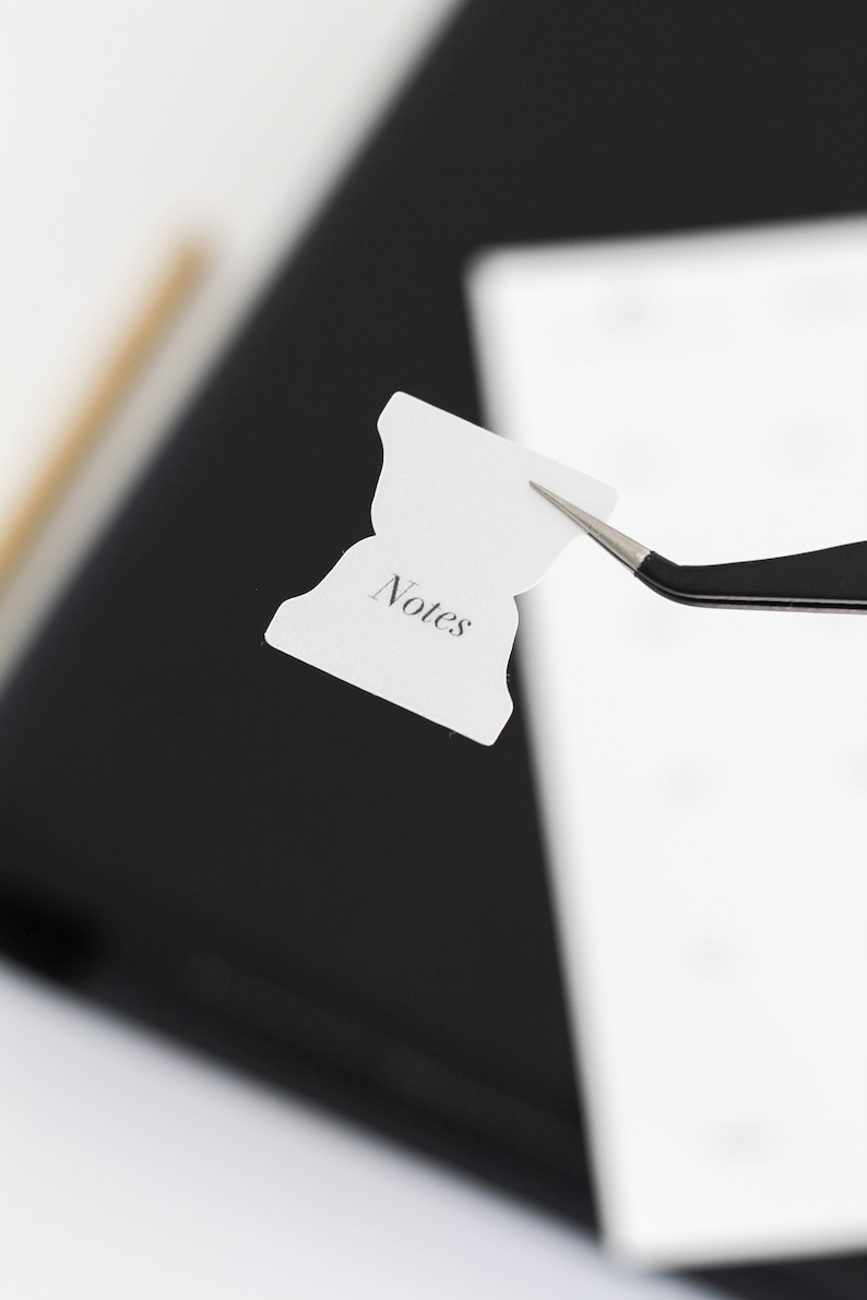 May include: A small white adhesive tab with a flared shape and the word Notes printed in a black serif font. The tab is held by a pair of black metal tweezers, showcasing its design for organization and labeling in planners or notebooks.