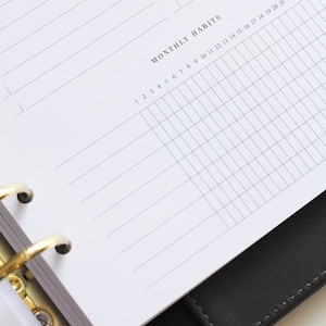 May include: A white planner page with black text featuring an 'IMPORTANT' section with horizontal lines and a 'MONTHLY HABITS' grid section numbered 1 through 27. The page is held by a gold-colored metal ring binder mechanism inside a black cover.