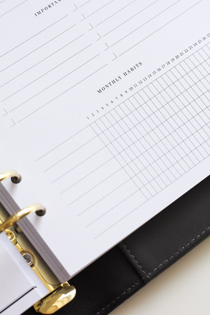 May include: A white planner page with black text featuring an 'IMPORTANT' section with horizontal lines and a 'MONTHLY HABITS' grid section numbered 1 through 27. The page is held by a gold-colored metal ring binder mechanism inside a black cover.
