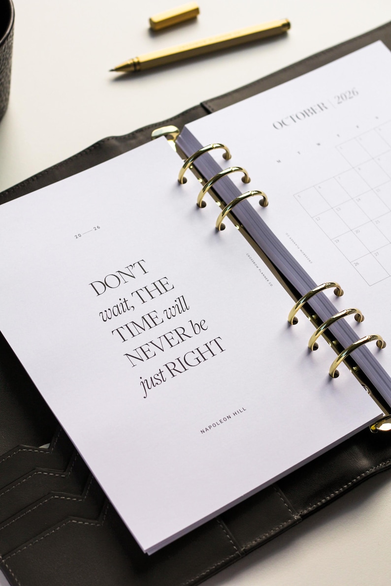 May include: Open planner with gold rings, featuring the quote "DON'T wait, THE TIME will NEVER be just RIGHT" by Napoleon Hill. The October 2026 calendar is visible. A gold pen rests nearby.