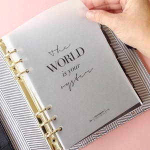 May include: A white translucent page with the text "the WORLD is your oyster" in black text. The page is in a black and white patterned binder with gold rings.