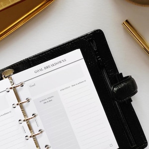 May include: A black leather bound planner with a white page open to a goal breakdown section. The page has headings for Goal, Strategy, Action Steps, and Achieve by. The planner has a zipper closure.