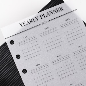 Peut inclure: Un agenda annuel pour 2026, avec des calendriers mensuels indiquant les dates et les jours de la semaine. L'agenda est imprimé sur du papier translucide et se trouve dans un classeur noir texturé. Le texte "YEARLY PLANNER" est en haut.