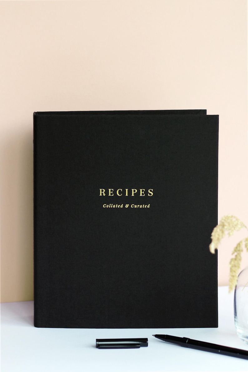 Puede incluir: Carpeta de recetas negra con letras doradas que dicen "RECIPES Collated & Curated".
