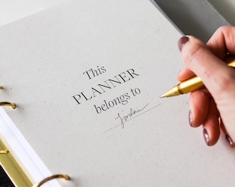 May include: A white planner with gold-toned metal rings and a gray elastic band. The page features the printed text 'This PLANNER belongs to' with the handwritten name 'Jordan' below it. A gold-toned pen is positioned on the page surface.