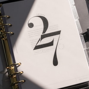 May include: A ring-bound planner with a gold-colored metal mechanism and black cover. The open page features the word TWENTY in small print above a large, stylized black number 27, with the words FOCUS and CONSISTENCY printed at the bottom.