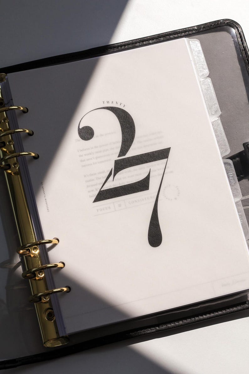 May include: A ring-bound planner with a gold-colored metal mechanism and black cover. The open page features the word TWENTY in small print above a large, stylized black number 27, with the words FOCUS and CONSISTENCY printed at the bottom.