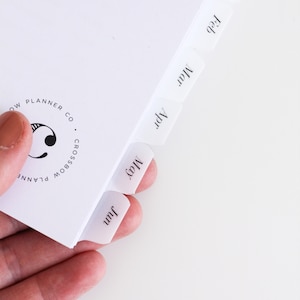 Könnte beinhalten: Ein weißer Planer mit monatlichen Registerkarten, die von Januar bis Dezember beschriftet sind. Der Planer hat ein kreisförmiges Logo mit der Aufschrift "Rainbow Planner Co."