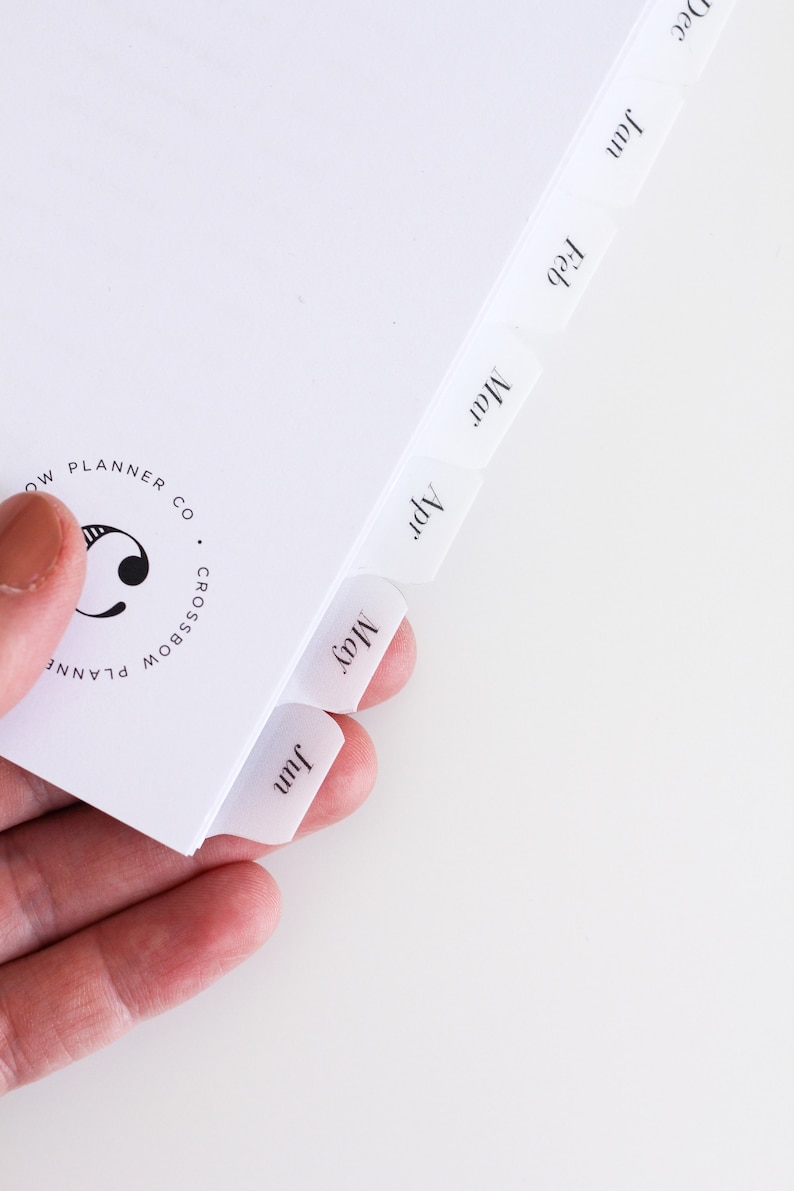 May include: A white planner page with monthly tabs labeled Jan, Feb, Mar, Apr, May, and Jun in a black serif font. The page features a circular logo with the text Crossbow Planner Co and a stylized letter C printed in black ink on the left side.